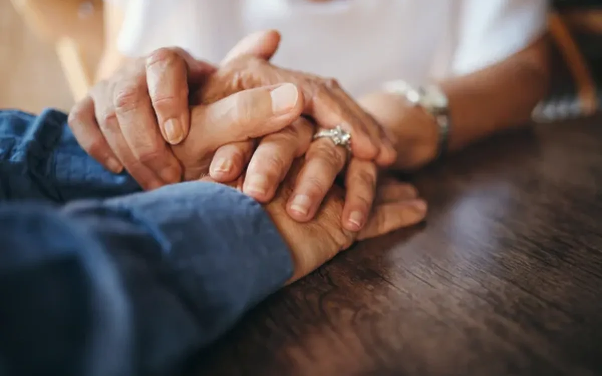 Mãos de uma pessoa idosa seguradas com carinho por outras mãos, sobre uma mesa de madeira