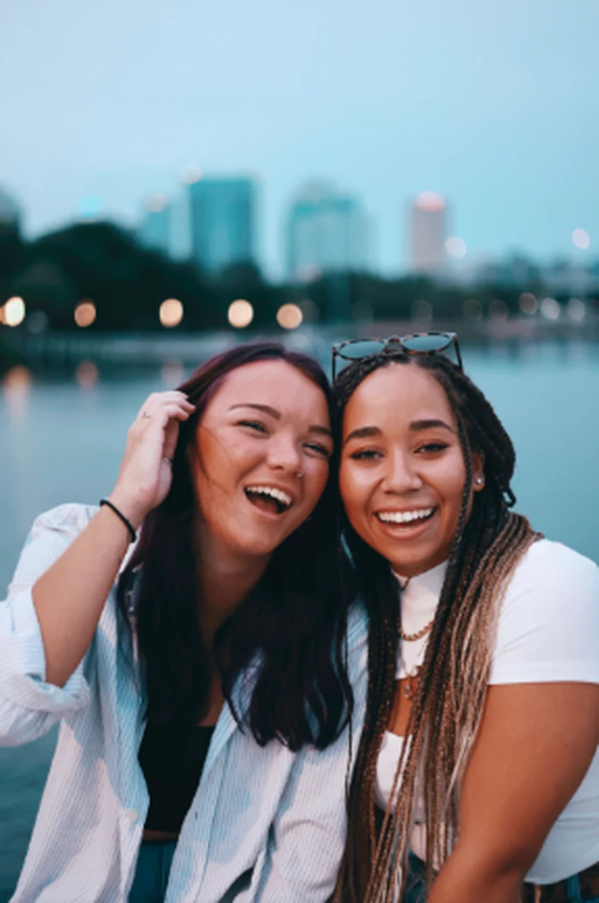 Duas jovens amigas rindo juntas à beira de um lago, com a cidade ao fundo