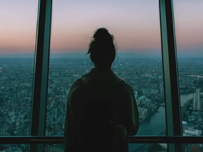 Mulher de costas olhando uma grande cidade ao entardecer por uma janela panorâmica
