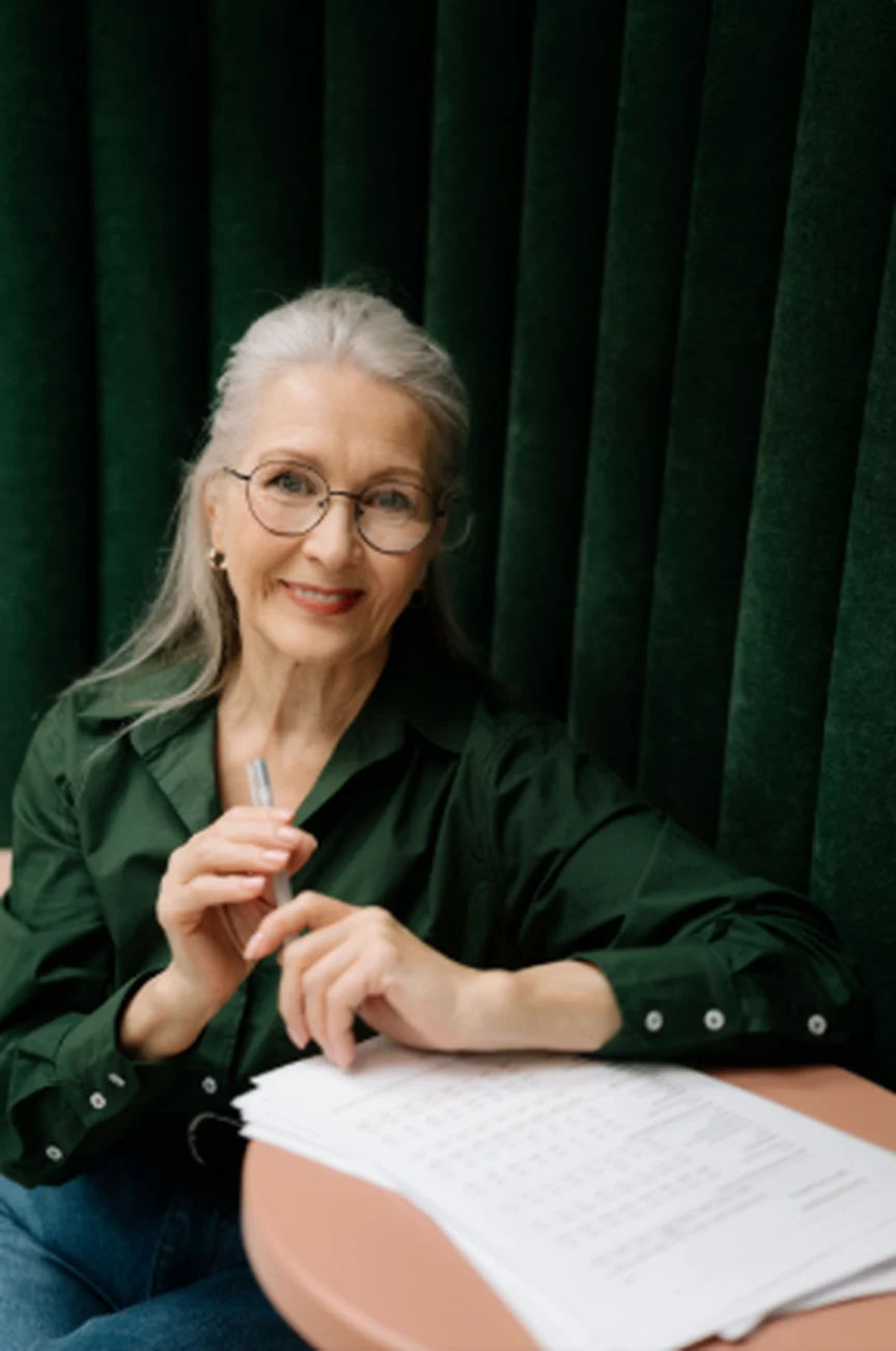 Mulher madura de cabelos grisalhos e óculos, sorrindo, sentada à mesa diante de uma cortina verde
